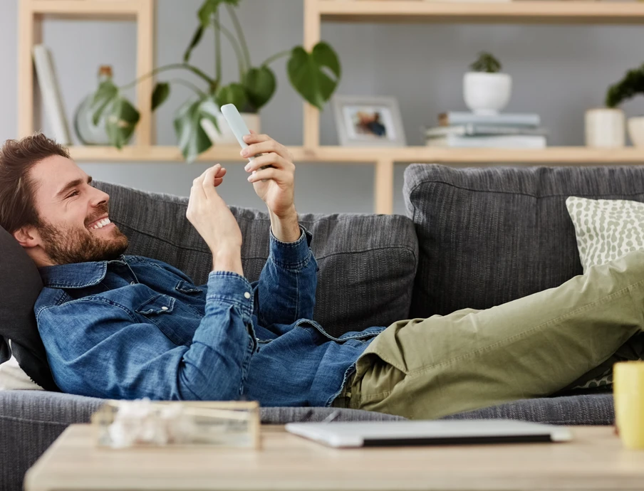Mann liegt entspannt auf einem grauen Sofa und schaut auf sein Smartphone; im Hintergrund Regale mit Pflanzen.