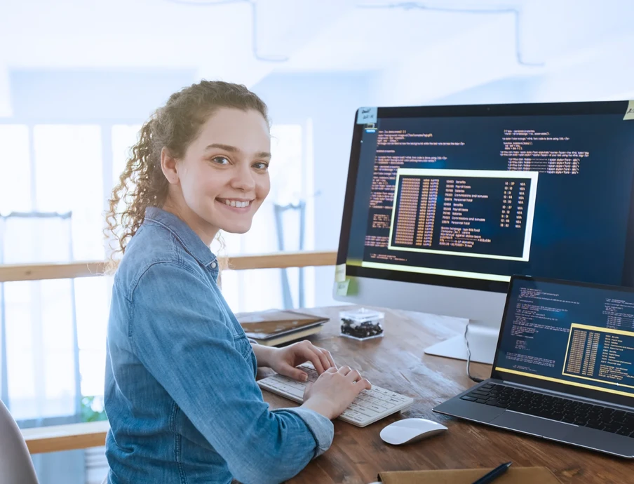 Frau mit Jeanshemd sitzt an Schreibtisch, Monitor und Laptop zeigen Code, sie lächelt.