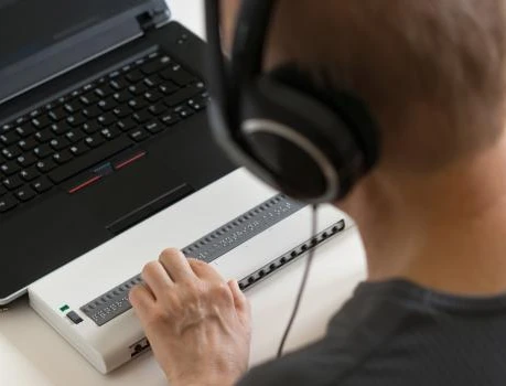 Person mit Kopfhörern vor Laptop; ein weißes Gerät mit vielen kleinen Tasten liegt auf dem Tisch.