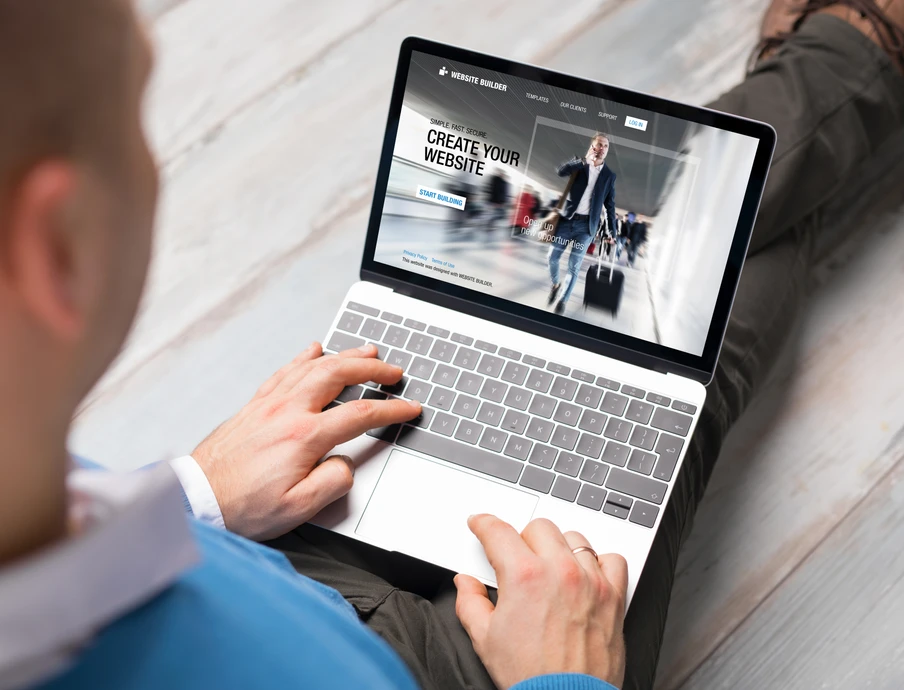 Person sitzt mit Laptop auf Holzboden; Laptopbildschirm zeigt Website-Entwurf mit dem Text Create Your Website.