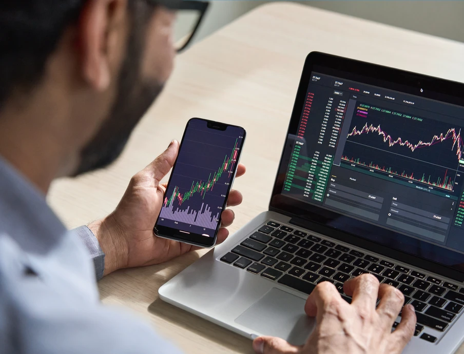 Person sitzt am Tisch, hält Smartphone mit Kursdiagramm; Laptop zeigt Börsencharts.