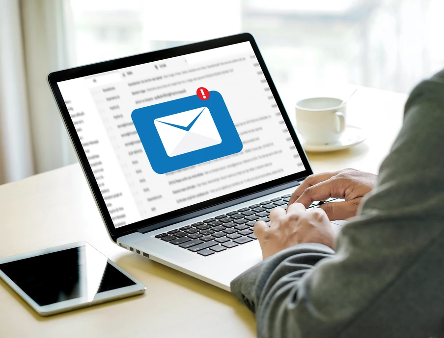 Person tippt am Laptop; Bildschirm zeigt blaues E-Mail-Symbol mit Benachrichtigung; daneben Smartphone und Kaffeetasse.