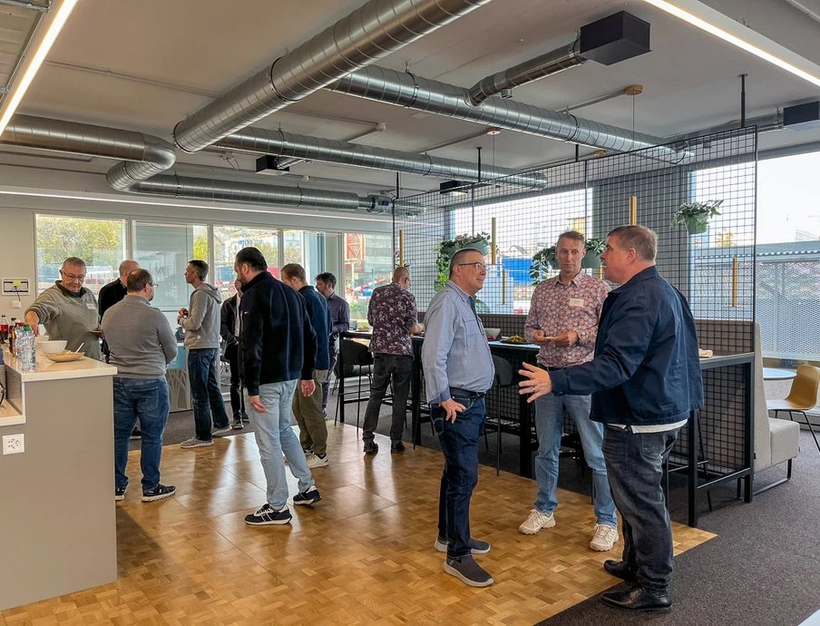 Gruppe von Menschen in einem Büro, stehend um hohe Tische; Lüftungsrohre, Holzboden, Glasfenster.