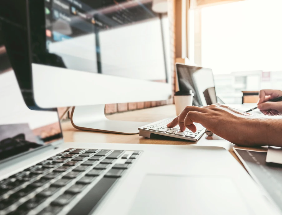 Schreibtisch mit Laptop, großem Monitor, Taschenrechner und Notizblock; Hände arbeiten an den Geräten.