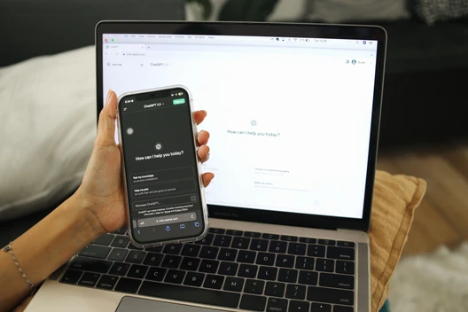 Eine Hand hält ein Smartphone vor einem geöffneten Laptop; Smartphone-Bildschirm zeigt Chat-Nachricht 'How can I help you today?'