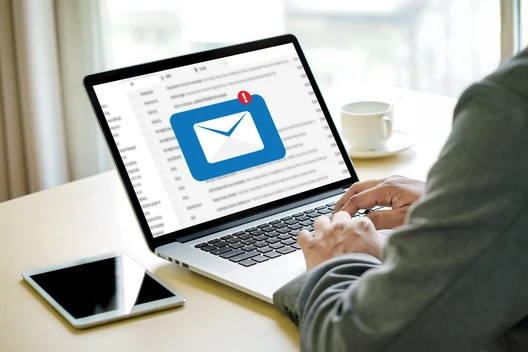 Person tippt am Laptop; Bildschirm zeigt blaues E-Mail-Symbol mit Benachrichtigung; daneben Smartphone und Kaffeetasse.