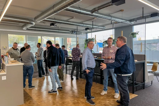 Gruppe von Menschen in einem Büro, stehend um hohe Tische; Lüftungsrohre, Holzboden, Glasfenster.