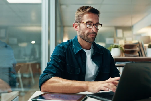 Mann mit Brille, dunkelblauem Hemd über weißem T-Shirt, sitzt am Schreibtisch und tippt auf Laptop; Tablet daneben.