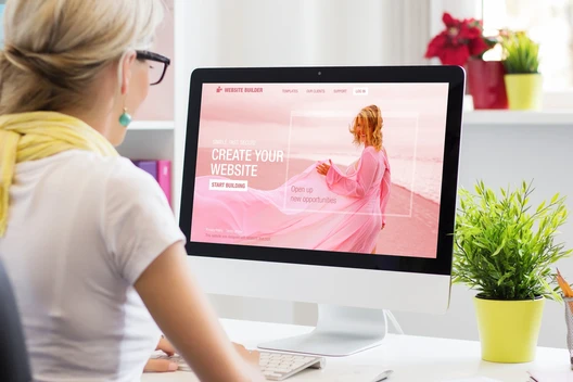 Person von hinten mit hellem Haar und Brille vor einem iMac; Bildschirm zeigt pinke Website, Schreibtisch mit Pflanze.